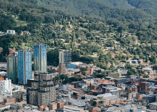 The city Bogota with modern high-rise buildings in shades of blue and green, set against a lush, forested hillside. The mix of contemporary and traditional architecture highlights the urban growth within a natural landscape.