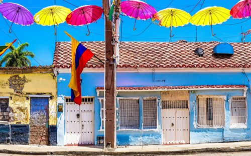 This is a nighttime cityscape of Cali, Colombia. The image features illuminated high-rise buildings nestled among green hills, with the city lights creating a warm glow. The iconic Cerro de las Tres Cruces can be seen in the background, adding to the city's distinctive skyline. The blend of urban and natural elements showcases Cali's vibrant atmosphere